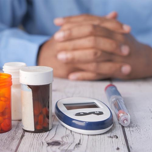 Bottles of pills, a syringe and a monitoring device sit on a table in front of a man's folded hands.