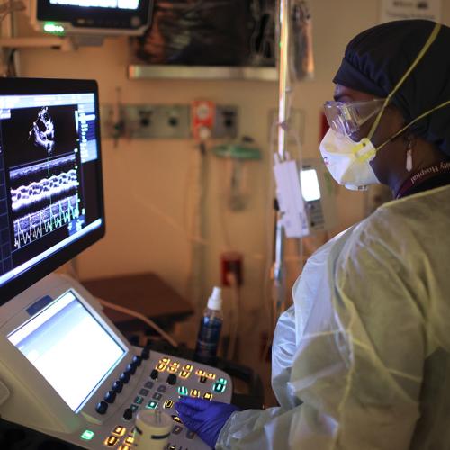 A masked health worker reads a screen. The screen shows a streaky image of a heart, surrounded by graphics and numbers.