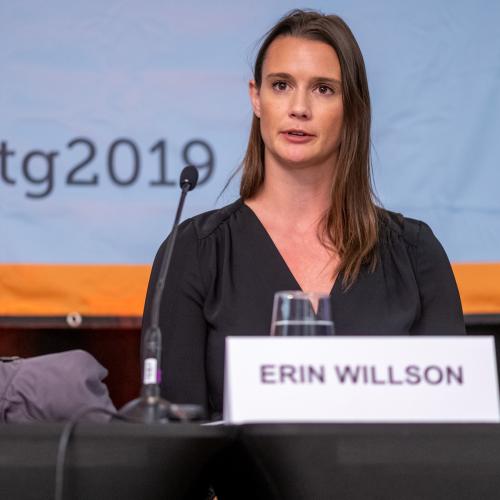 Erin Willson speaks as she sits at a conference table. On the screen behind is the hashtag #ptg2019. 