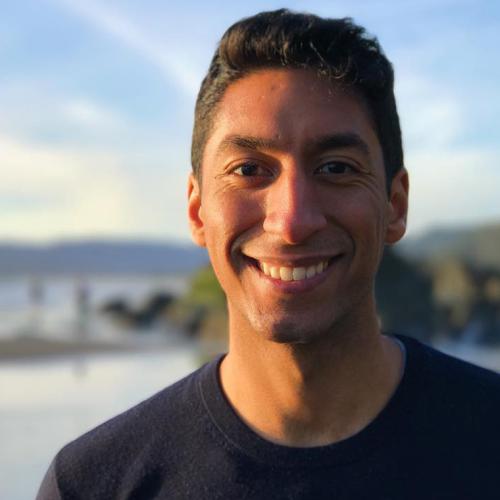 Zain Manji smiles, standing in front of a wooded lake at sunset.
