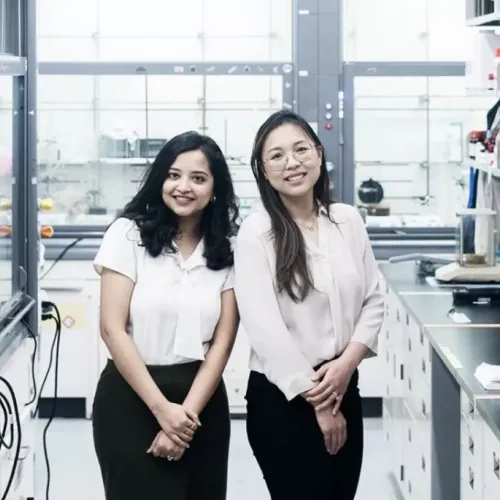 Scientists Nabanita Nawar and Pimyupa Manaswiyoungkul, both wearing white blouses and black pants, stand together in their lab smiling.