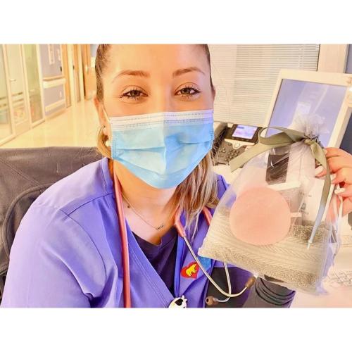 A young woman wearing a mask and scrubs holds up a bag containing a towel and a tube of a skin-care cream.