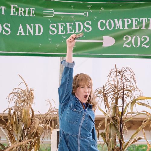 A woman in a denim jumpsuit standing with one arm overhead in front of some dried corn stalks and a sign that reads Fort Erie Weeds and Seeds Competition 2025