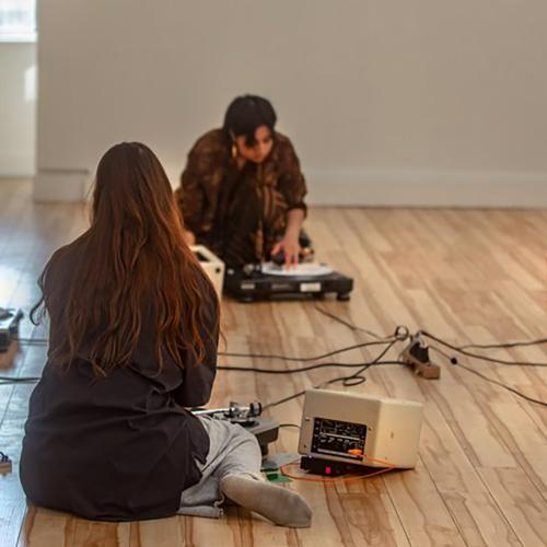 A group of young people sitting on a hardwood floor with some music equipment