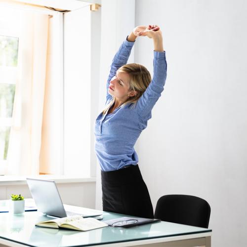 A woman stands beside a desk, stretching up with her arms above her head.