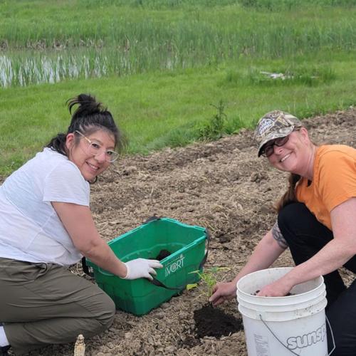 Lisa Owl and Melanie Jeffrey smile as they kneel and place a crookneck squash seedling in the earth.