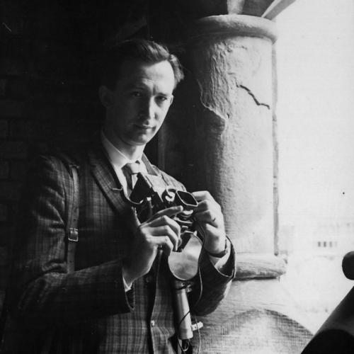 Robert Lansdale stands in shadow on a stone balcony, looking out at the light and holding a camera.