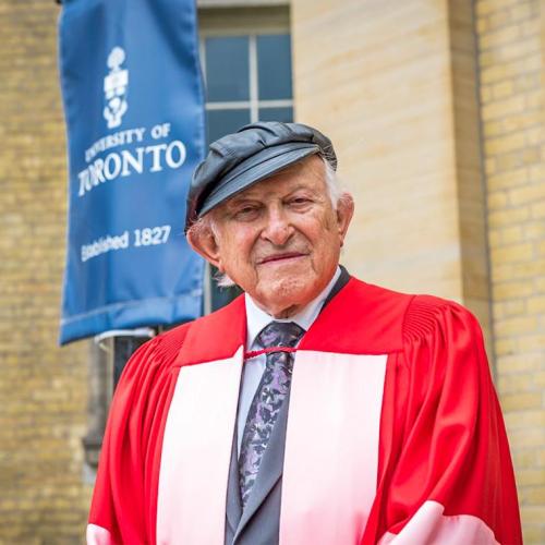 Nate Leipziger, wearing academic robes and his trademark leather cap, stands outside Convocation Hall.