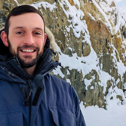 Luke Spooner smiles, as he stands in front of a snow-covered cliff, in a spruce forest.