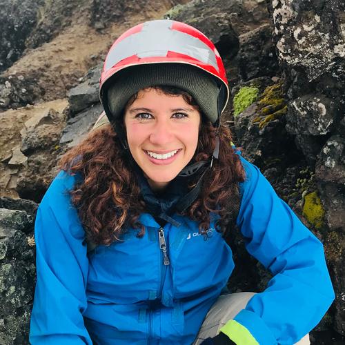 Marah Ayad smiles. She is wearing a hard hat and hiking gear and standing in front of a rocky cliff face.