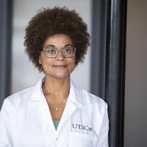 Maydianne Andrare smiles and looks thoughtful, standing in a hallway and wearing a UTSC lab coat.