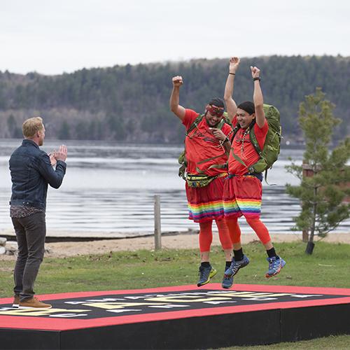 Anthony Johnson and James Makokis, wearing matching T-shirts and rainbow skirts, jump for joy on the winner's mat of Amazing Race Canada.