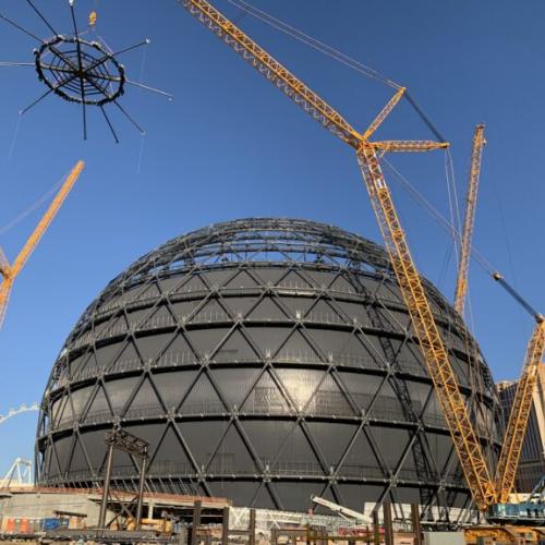 A crane lifts a spiderweb-shaped object high above a huge sphere built out of triangles.