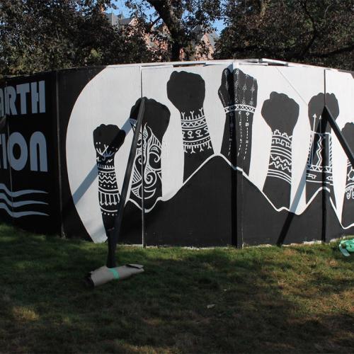 Plywood panels circle a tree. They have been painted with an image of tattooed arms raising their fists in the air.