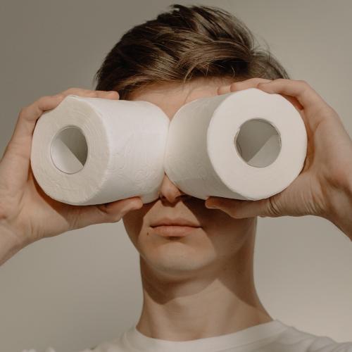 A young man holds rolls of toilet paper up to his eyes as if they were binoculars.