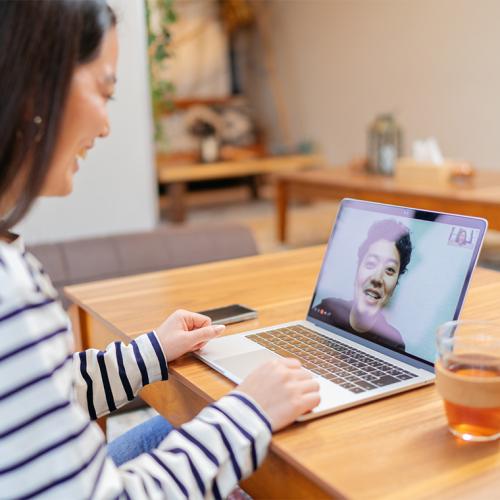 Two women laugh as they talk in a video call, one sitting looking at a laptop screen and the other on the screen itself.