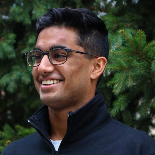 Shrey Jain laughs while standing near fir trees outdoors.