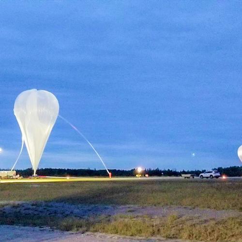 Two giant balloons, anchored in a field at dusk, gradually swell with hot air. The gondolas beneath are too small to see.