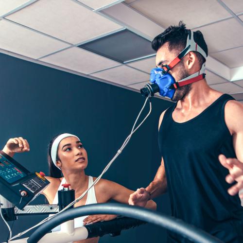 A young man runs on a treadmill, wearing a mask to measure his breathing. A young woman operates the controls.