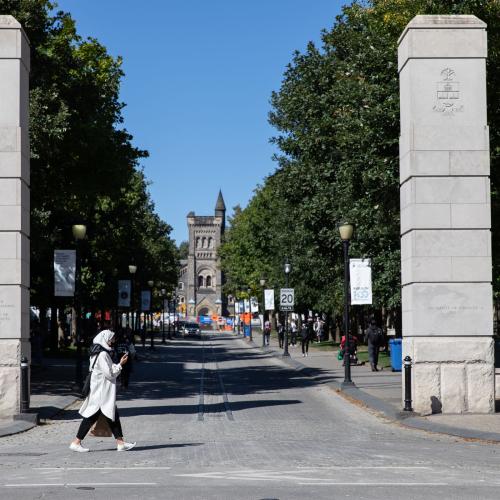 A woman walks past two stone pillars flanking a cobbled street. University College can be seen at the end of the street.