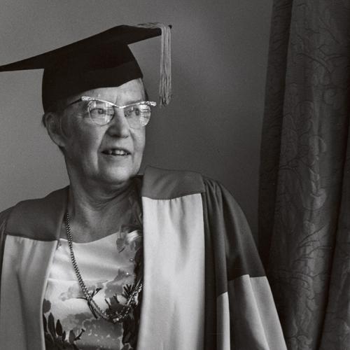 In a historical photograph, Elsie McGill stands by a window, wearing her academic hat and robes.