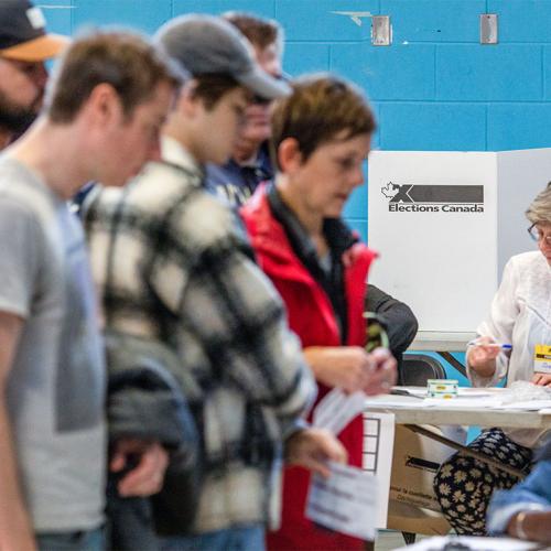 Several people line up at a polling station in a gymnasium as a volunteer sits in front of the cardboard polling booth.