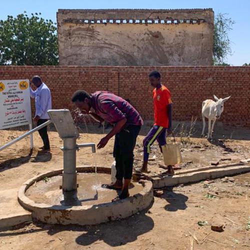 Beside a dusty road, a boy works a metal pump handle while a man drinks.