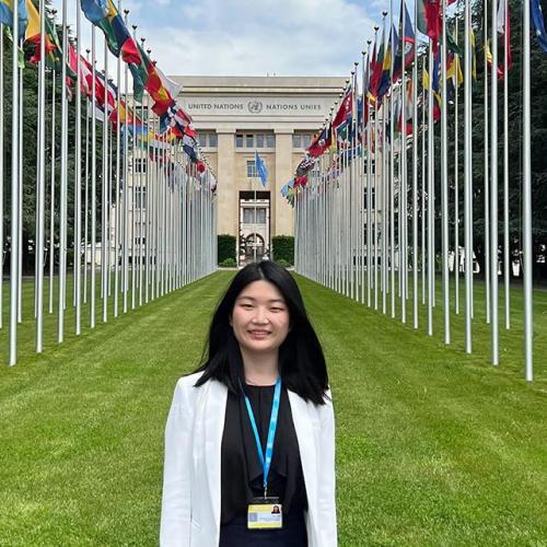 Yifan Zhou smiling. standing next to the rows of flags outside the United Nations building in Geneva.