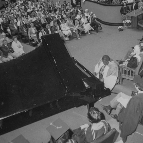 Oscar Peterson plays a grand piano on stage at Convocation Hall, surrounded by professors in academic robes.