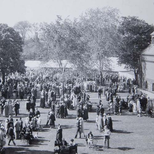 In June 1926, hundreds of people in formal clothes and hats chat in groups on a lawn by a tent and trees.