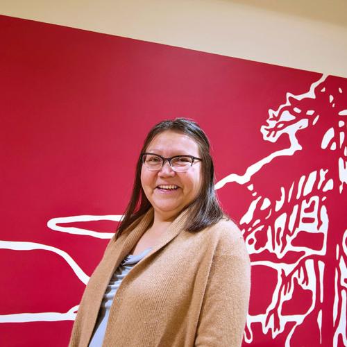 Assistant Professor Brenda Wastasecoot (PhD 2017) smiling in front of artwork of a pine tree (photo by Diana Tyszko) 