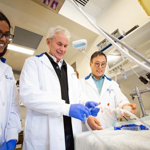Arthur Slutsky, wearing gloves and a lab coat, shows a plastic tube with valves to colleagues (photo by Roberta Baker)