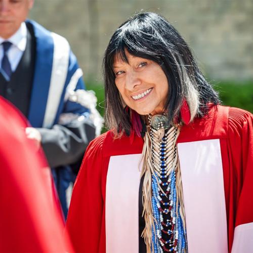 Buffy Sainte-Marie smiles mischievously as she looks through a crowd of dons, wearing academic robes.