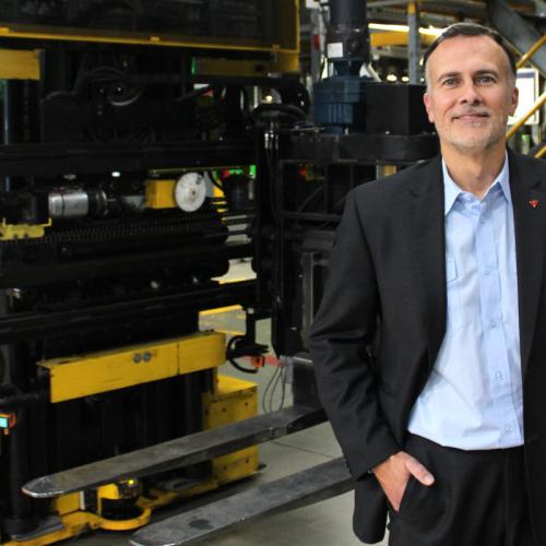 Carlos Menezes smiles as he stands in front of a forklift in a warehouse with one hand in his pocket.