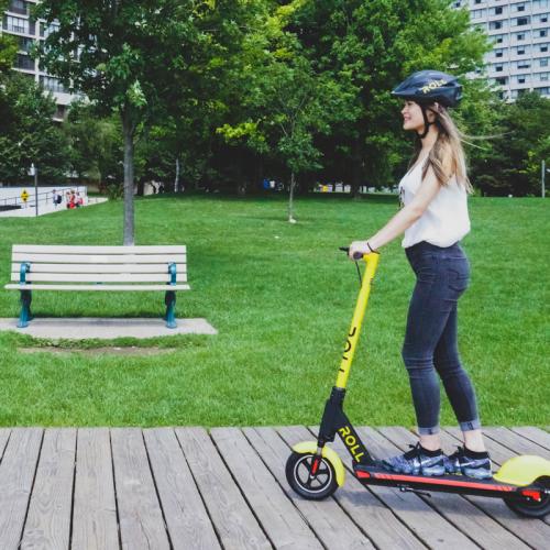 A woman wearing a helmet stands on a Roll brand e-scooter as she rolls along a boardwalk past a park bench.