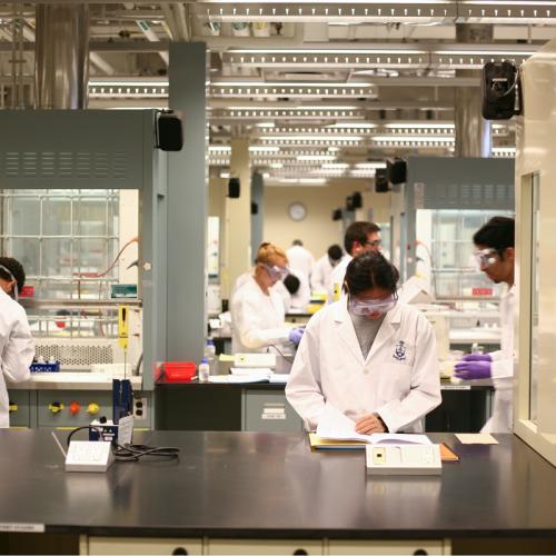 A diverse group of people in lab coats and safety goggles work intently the beakers, books and equipment in a lab at U of T's Department of Chemistry.