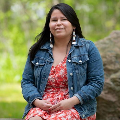 Diane Hill smiles, sitting on a rock in a meadow outdoors.