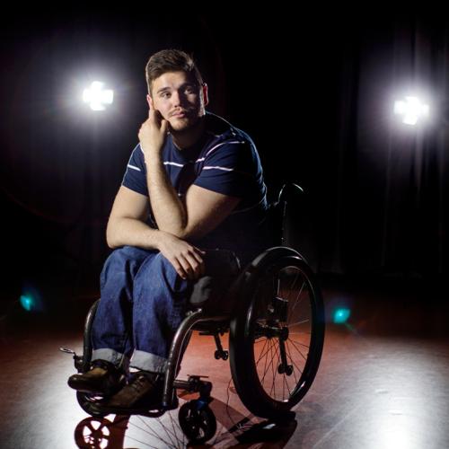 George Alevizos sits in his wheelchair, looking thoughtful, in the spotlight on a bare stage.