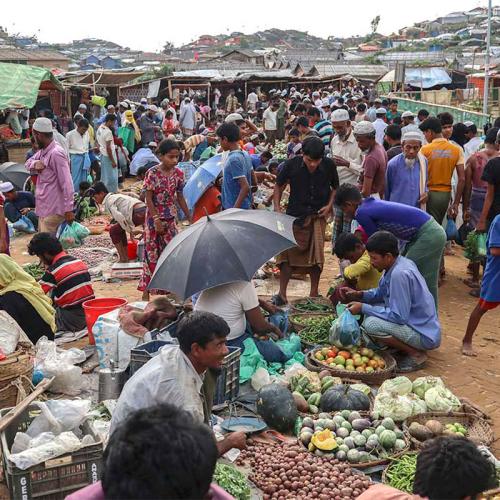 People walk through a crowded, open-air market, between roughly-built shacks, as vendors display food in baskets and sacks.