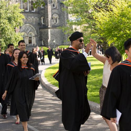 A student in academic robes steps out of line to high five a friend as graduates walk along King's College Circle to Convocation Hall.