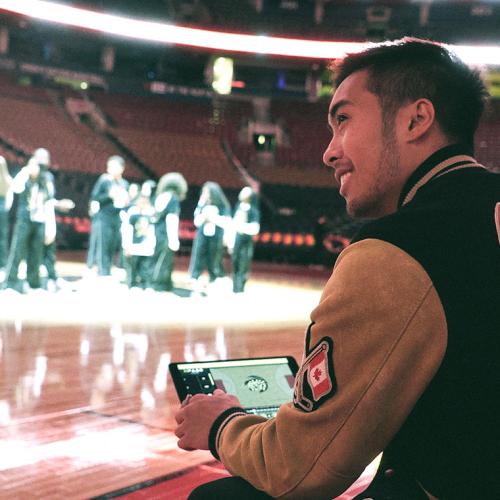 Axel Villamil sits in the foreground holding a tablet with his app and watching the Raptors dancers gather on the court in the background (photo courtesy of StageKeep) 