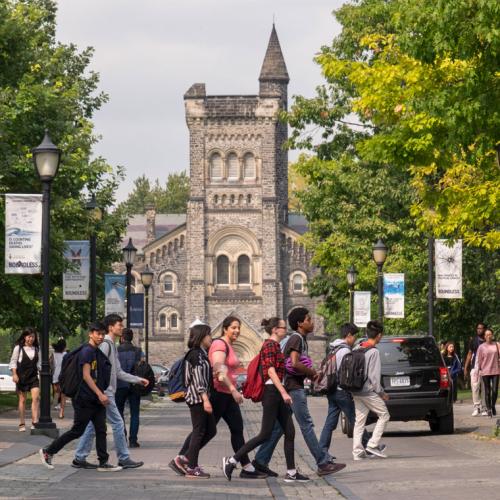 A crowd of chatting students cross the road while behind them, University College peeks through the trees 