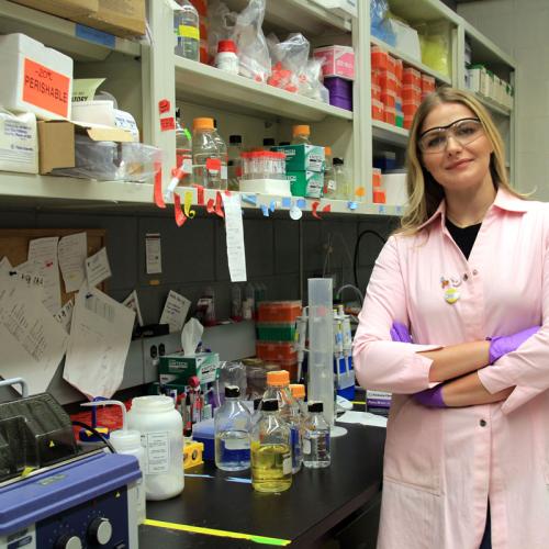 Sasha Weiditch, in lab coat and safety goggles, next to shelves crowded with lab gear, chemicals, sticky notes and posters.