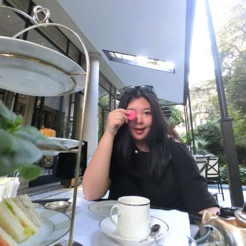 Joanna Luo smiles and holds a pink macaron cookie over one eye, while sitting at an outdoor cafe.
