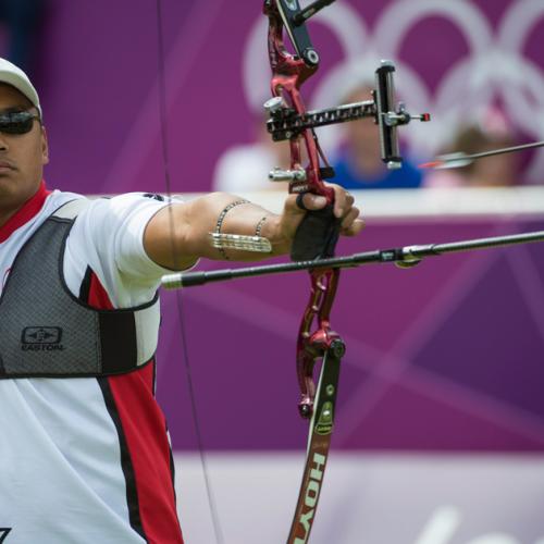Crispin Duenas releases an arrow from his bow. The Olympic rings are seen on a wall in the distance.