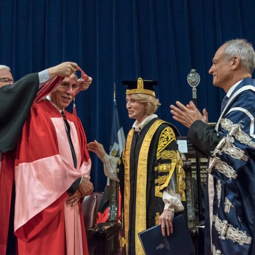 A don lifts the academic hood over Alan Bernstein's head as Chancellor Patten and President Gertler applaud.