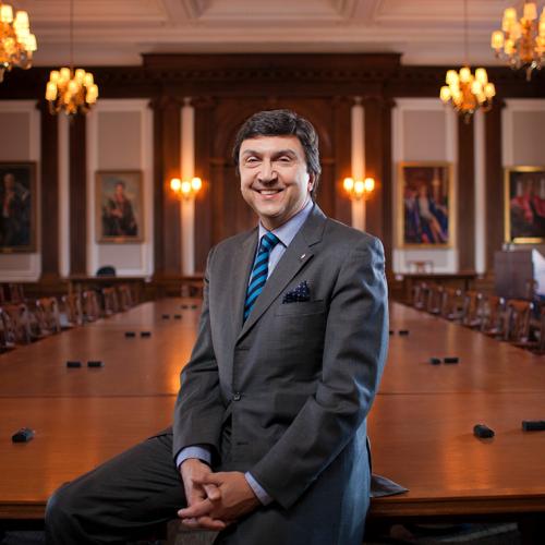 David Naylor smiles while leaning on a  polished conference table in a large boardroom, decorated with painted portraits and old-fashioned chairs.
