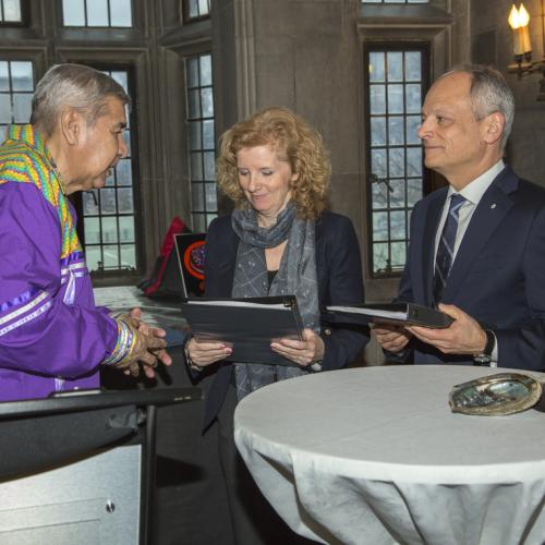 Elder Andrew Wesley, wearing formal Indigenous regalia, hands binders to Cheryl Regehr and Meric Gertler by a table holding a sacred abalone shell (photo by Johnny Guatto)