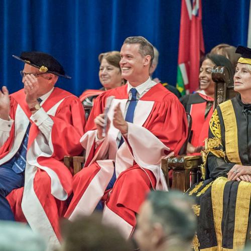 Mark Tewksbury grins and applauds while sitting on stage at Convocation Hall with Rose Patten, wearing academic robes.