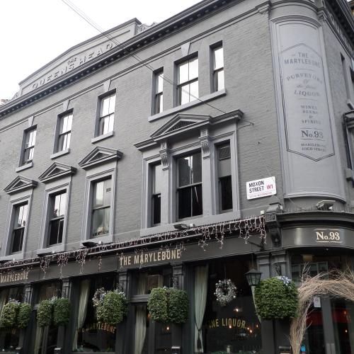 An exterior photo of the Marylebone Pub, London. It's a grey building on the corner of Marylebone High street and Moxon Street. The former name of the pub is visible at the top, etched in the stone (The Queen's Head)